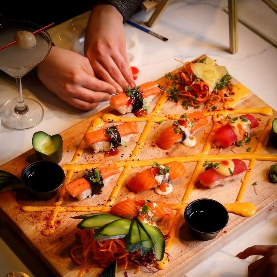 Hands arranging sushi on a wooden board with a cocktail and painting supplies nearby.
