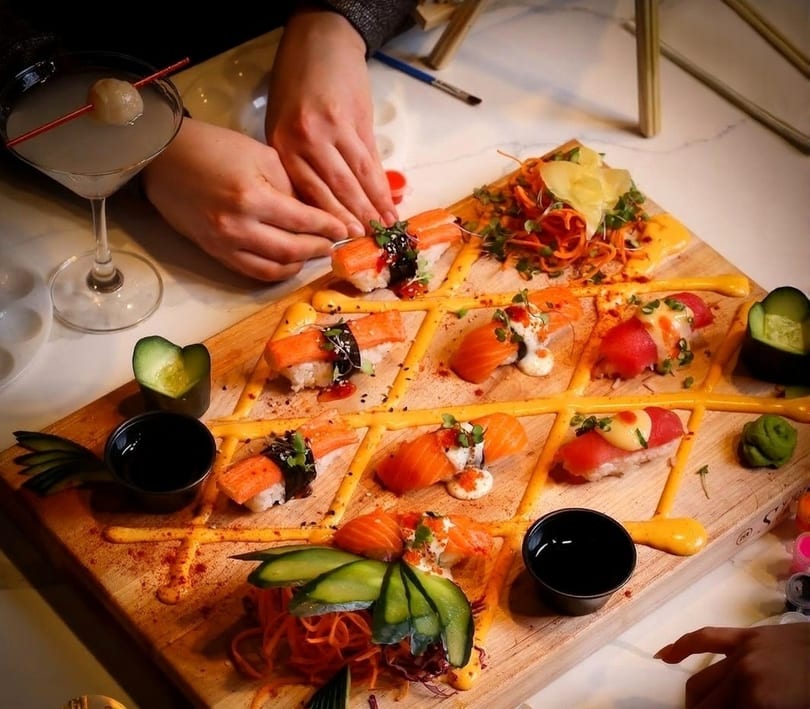Hands arranging sushi on a wooden board with a cocktail and painting supplies nearby.