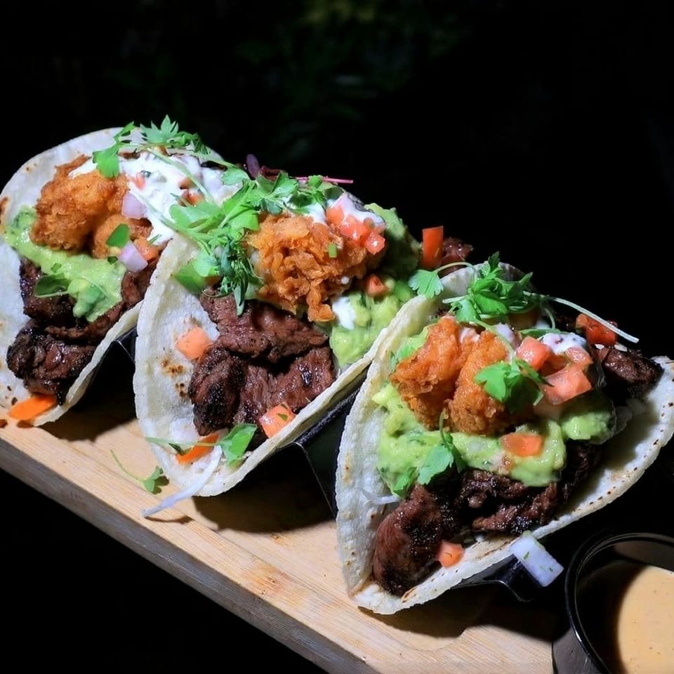 Three gourmet tacos with steak, guacamole, fried shrimp, and pico de gallo on a wooden board.
