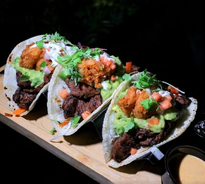Three gourmet tacos with steak, guacamole, fried shrimp, and pico de gallo on a wooden board.